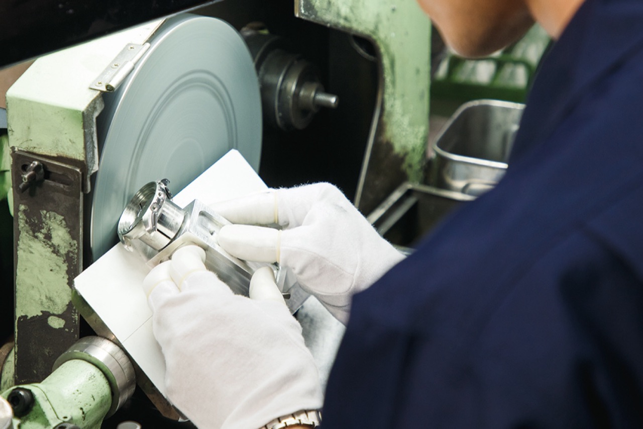 A craftsperson in white cotton gloves pressing a clamped watch case against the face of a large rotating flat disc on a pale green polishing machine