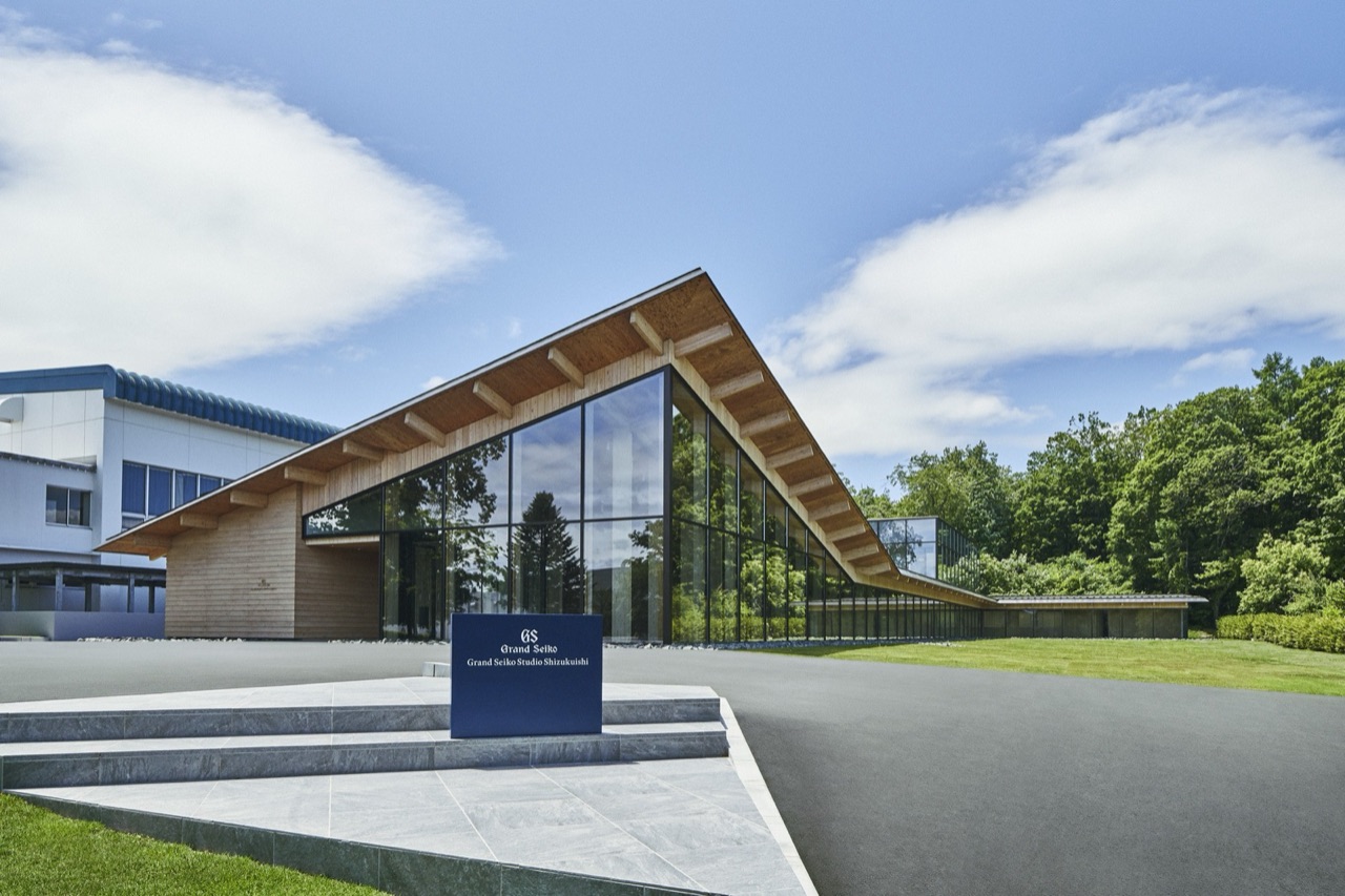 The Grand Seiko Studio Shizukuishi, a low building under a peaked roof of exposed timber beams above a fully glazed wall, set against summer woodland, with a dark navy sign on a stone plinth in the foreground